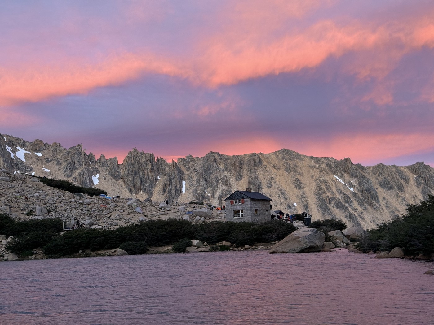 Refugio Frey beneath jagged mountain peaks and a pink sunset sky, with a lake in the foreground